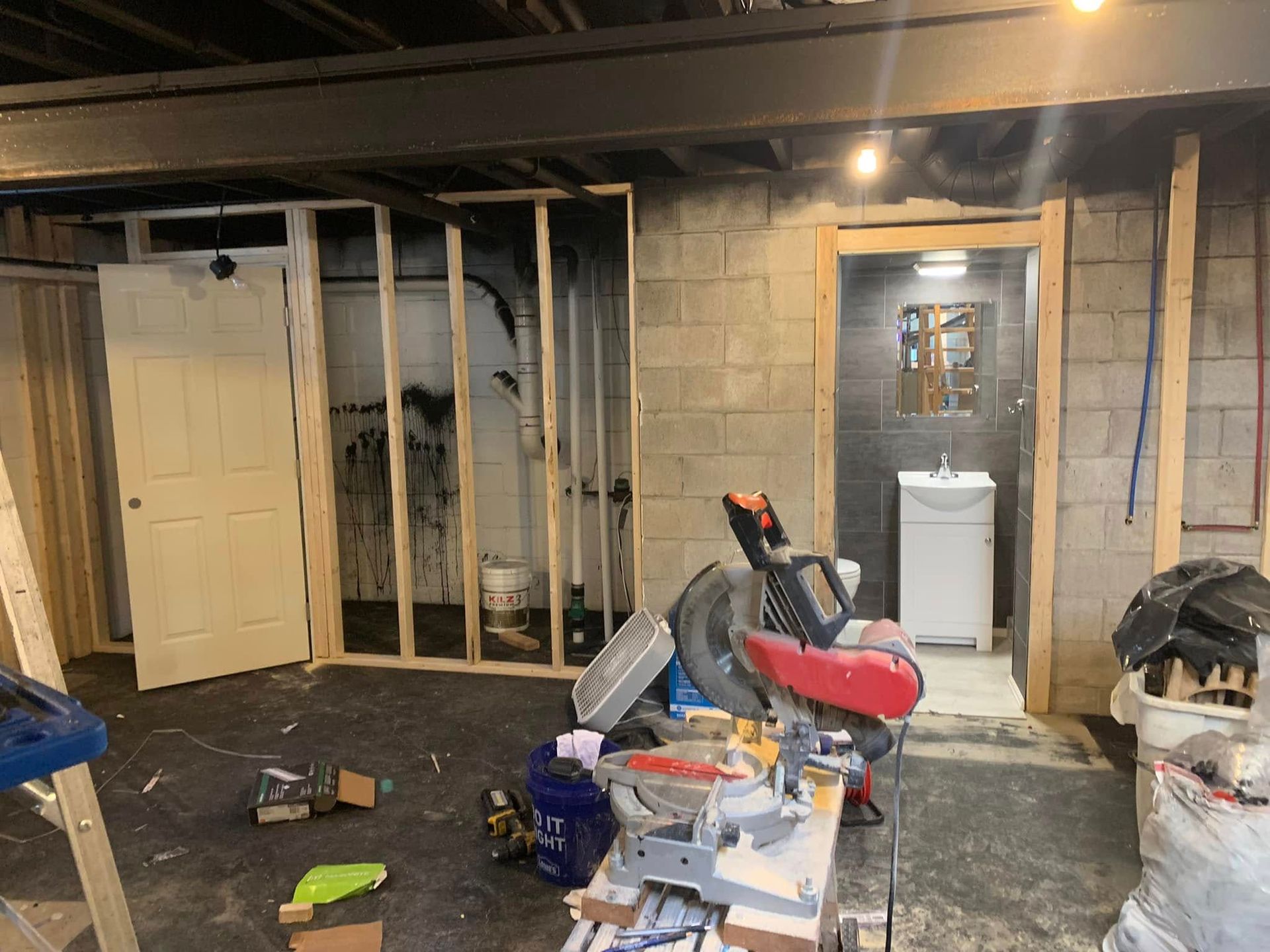 A basement under construction with wooden wall framing, a door, a miter saw on a table, and a finished bathroom beyond.