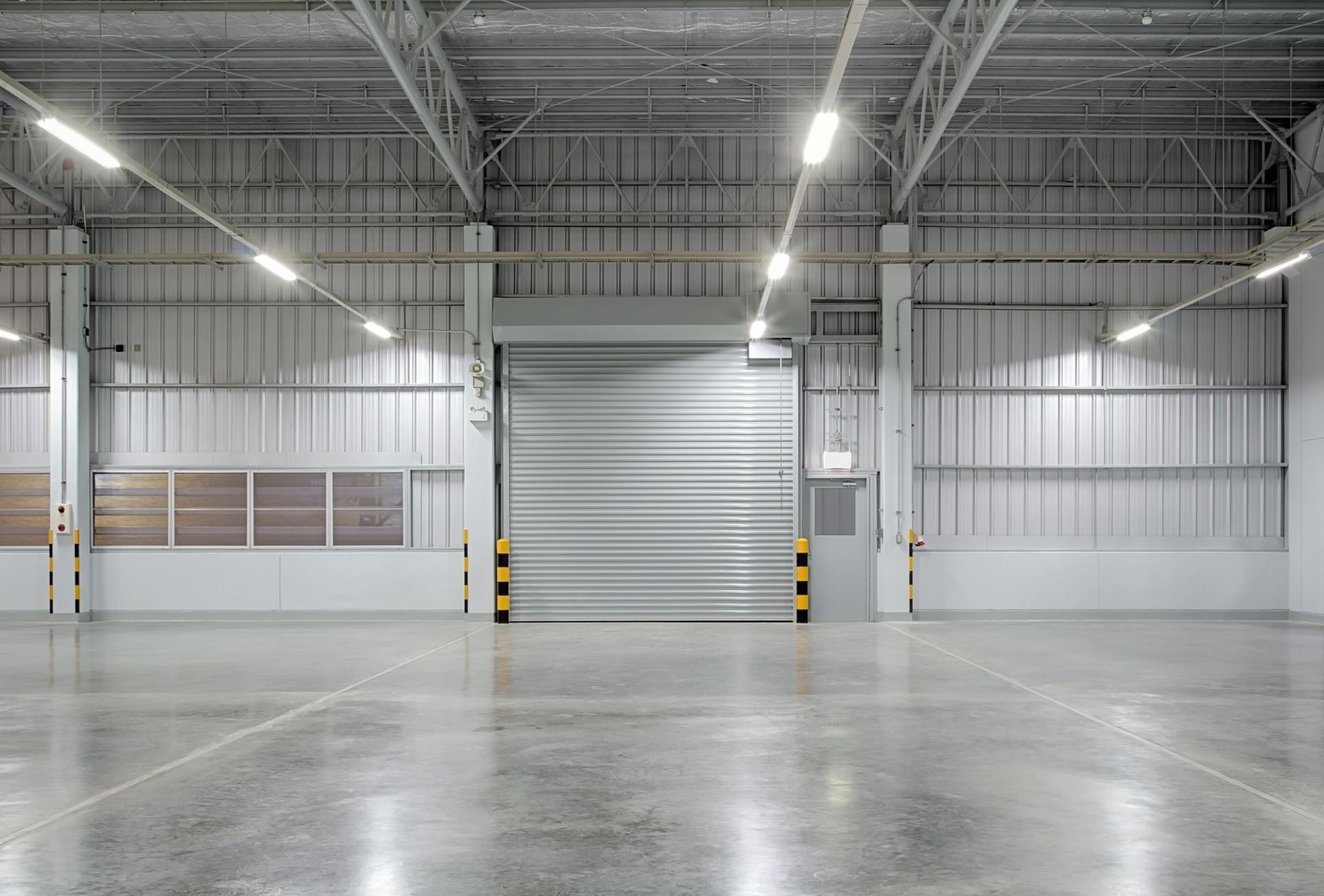 An empty industrial warehouse with a large grey rolling shutter door and polished concrete floors under bright lighting.