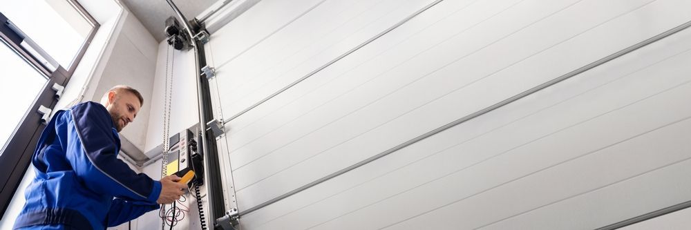 A worker in a hard hat and protective clothing installs or repairs the vertical track of a white sectional garage door.