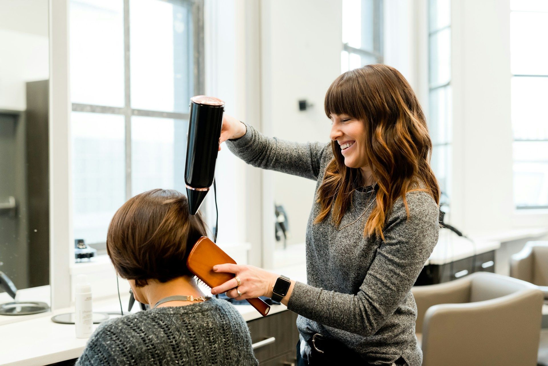 Hairdresser styling a client's hair in a salon; smiling, using a hair dryer and brush.