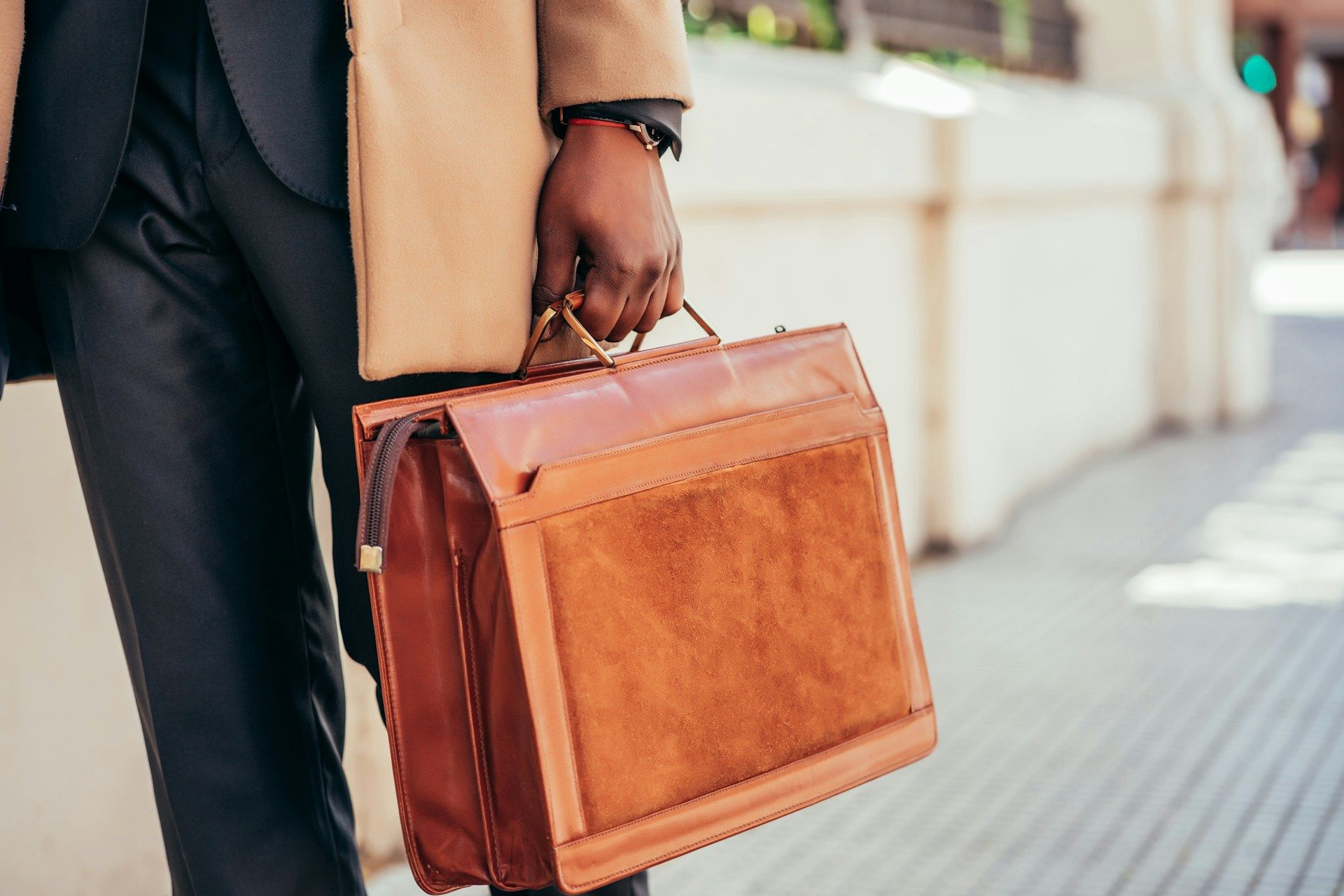 Person in suit holding a brown briefcase outdoors, near a building.