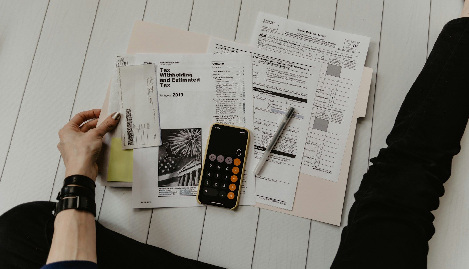 Person sitting on the floor reviews documents, calculator, and pen.  Black pants, watch, and pale wood floor.
