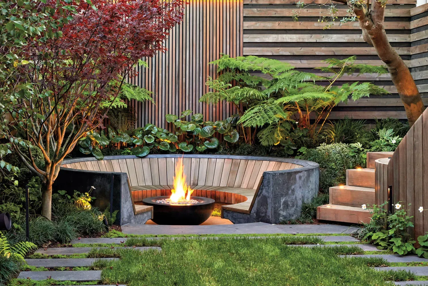 Old Bridge Landscape Contractor. Modern backyard fire pit surrounded by stone benches, greenery, and wooden fencing at dusk