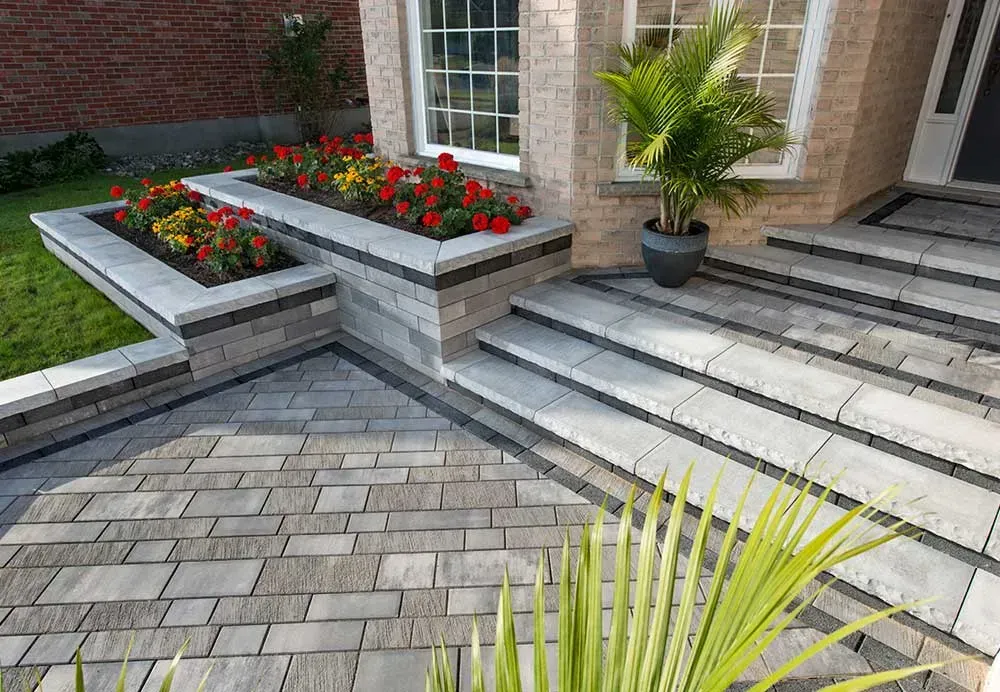 Morganville Landscaper. Modern stone patio with raised flower beds, red blooms, and potted palm beside brick house steps