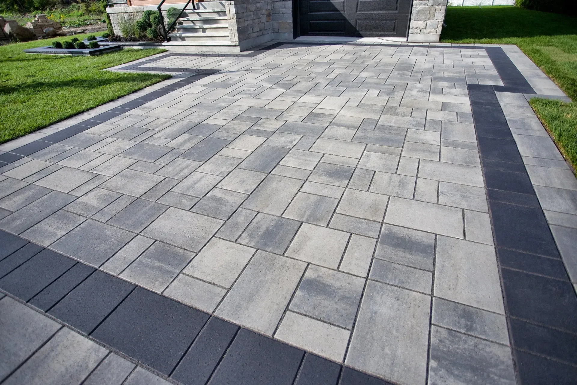 Paved stone walkway in Colts Neck with a gray and light tan pattern beside a lawn and building entrance