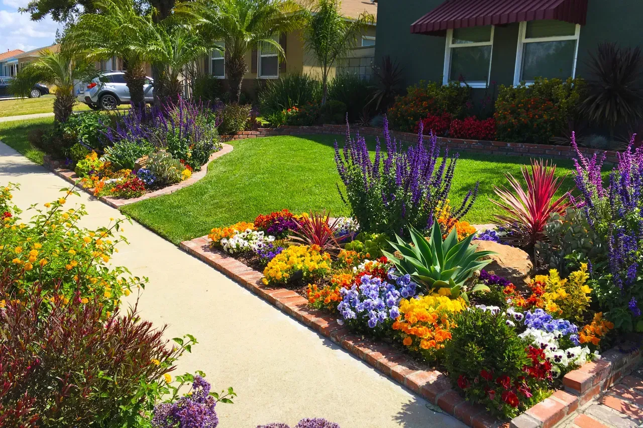 Colorful flower beds line a sunlit sidewalk beside a suburban house and manicured lawn. Landscaping for Old Bridge NJ
