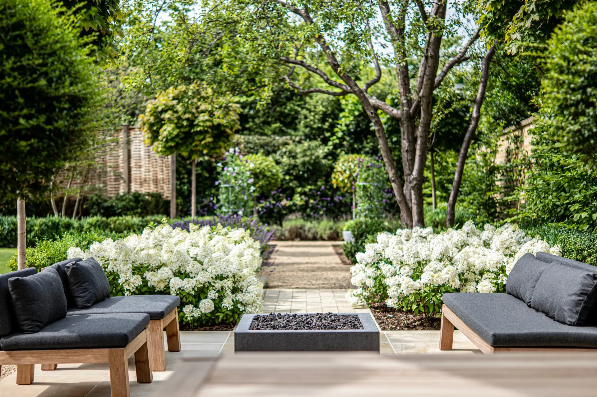 Landscape Construction Atlantic Highlands NJ. Modern garden patio with two cushioned benches, a fire pit, and white flowers beneath green trees