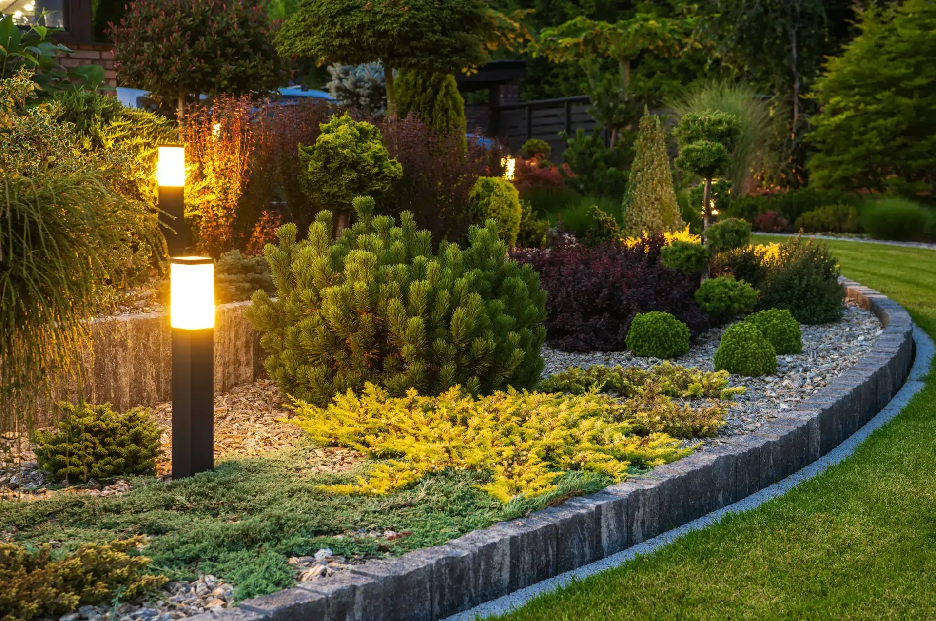 Landscape Lighting Company in Monmouth County Illuminated landscaped garden with shrubs, flowers, and a curved stone border at dusk