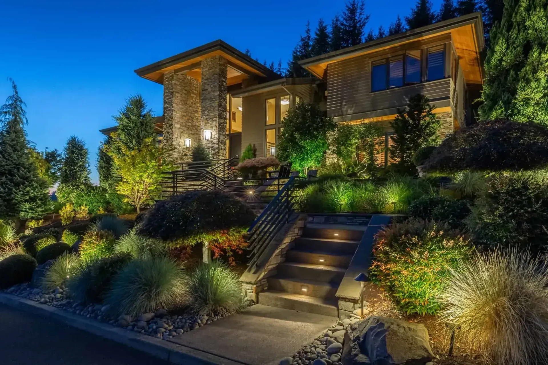 Landscape lighting in Holmdel. Luxury hillside home at dusk with lit stone steps, landscaped garden, and warm glowing windows