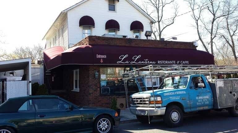 Blue tow truck parked outside a brick storefront with a maroon awning; a dark car is parked beside it.