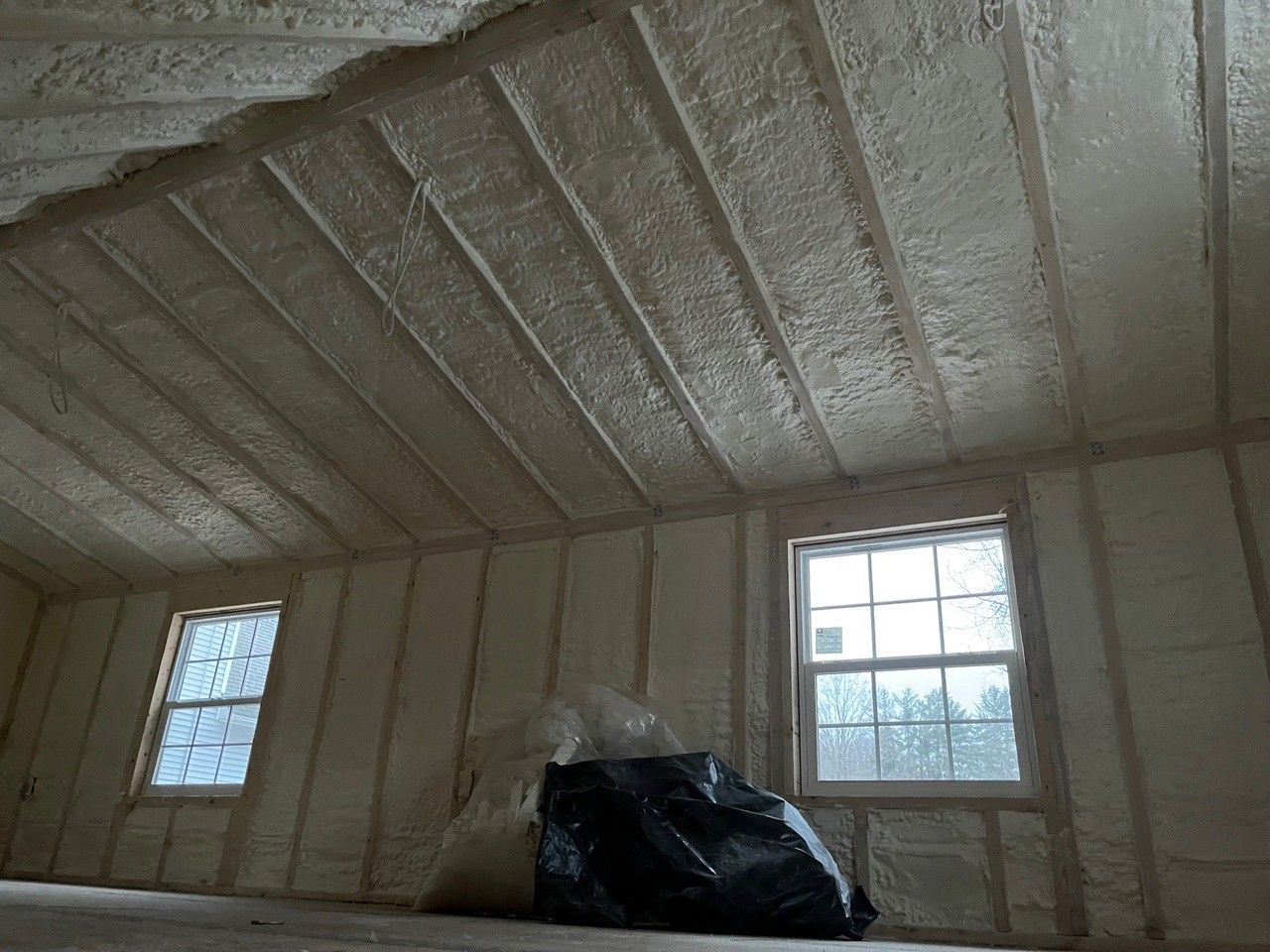 Attic space insulated with beige foam. Windows along wall, beams and framing visible. Black bag in front.
