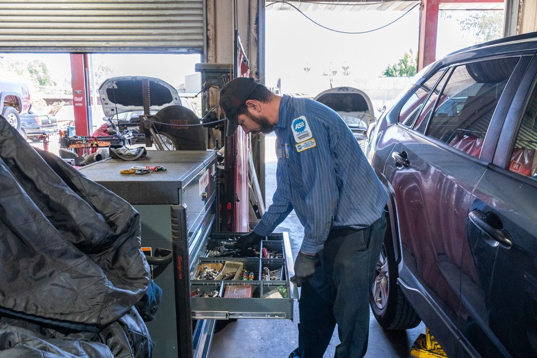 Mechanic working on a car in a garage beside a purple SUV, with tools and parts nearby