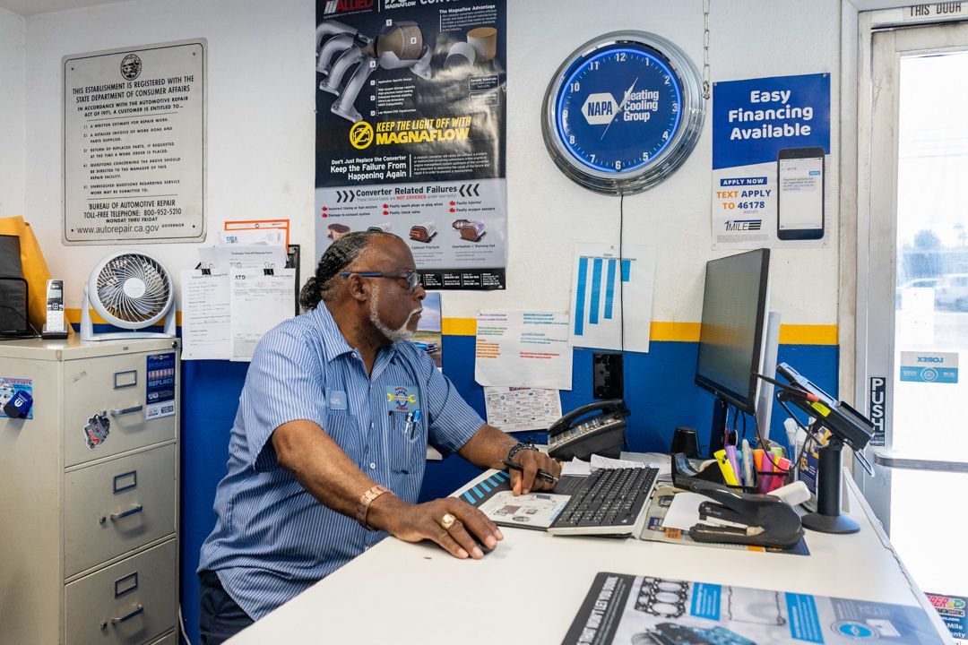 Person seated at an office desk with computers, phone, and wall posters in a small workspace