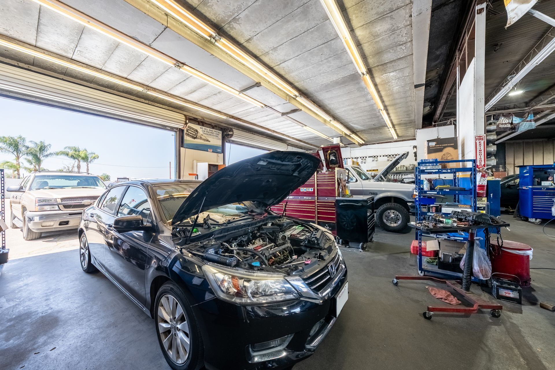 Black car with open hood in an auto repair shop, mechanic working nearby and tools on a cart.