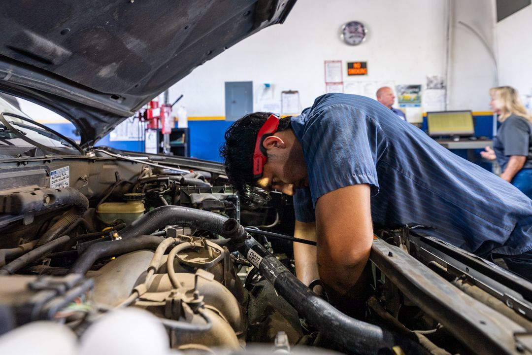 Mechanic leaning into an open car engine bay in a garage, wearing blue coveralls and red earmuffs.