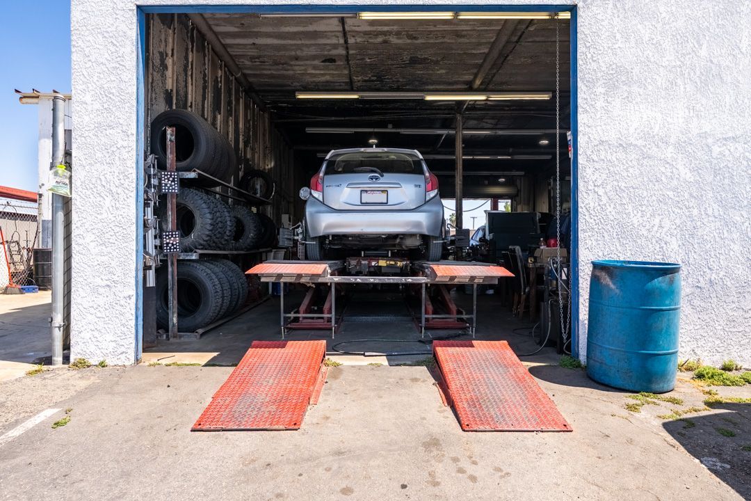 Car in an auto repair garage on ramps, with tires stacked on the left and a blue barrel on the right