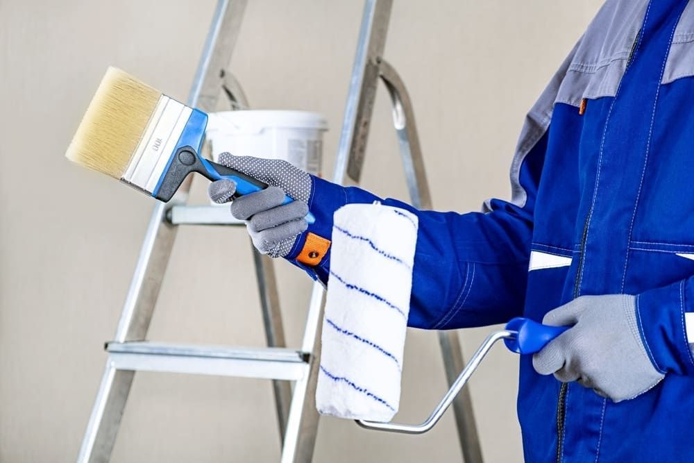 A Man Is Holding A Paint Roller And A Brush In Front Of A Ladder — Ballina Painting Service In Lennox Head, NSW