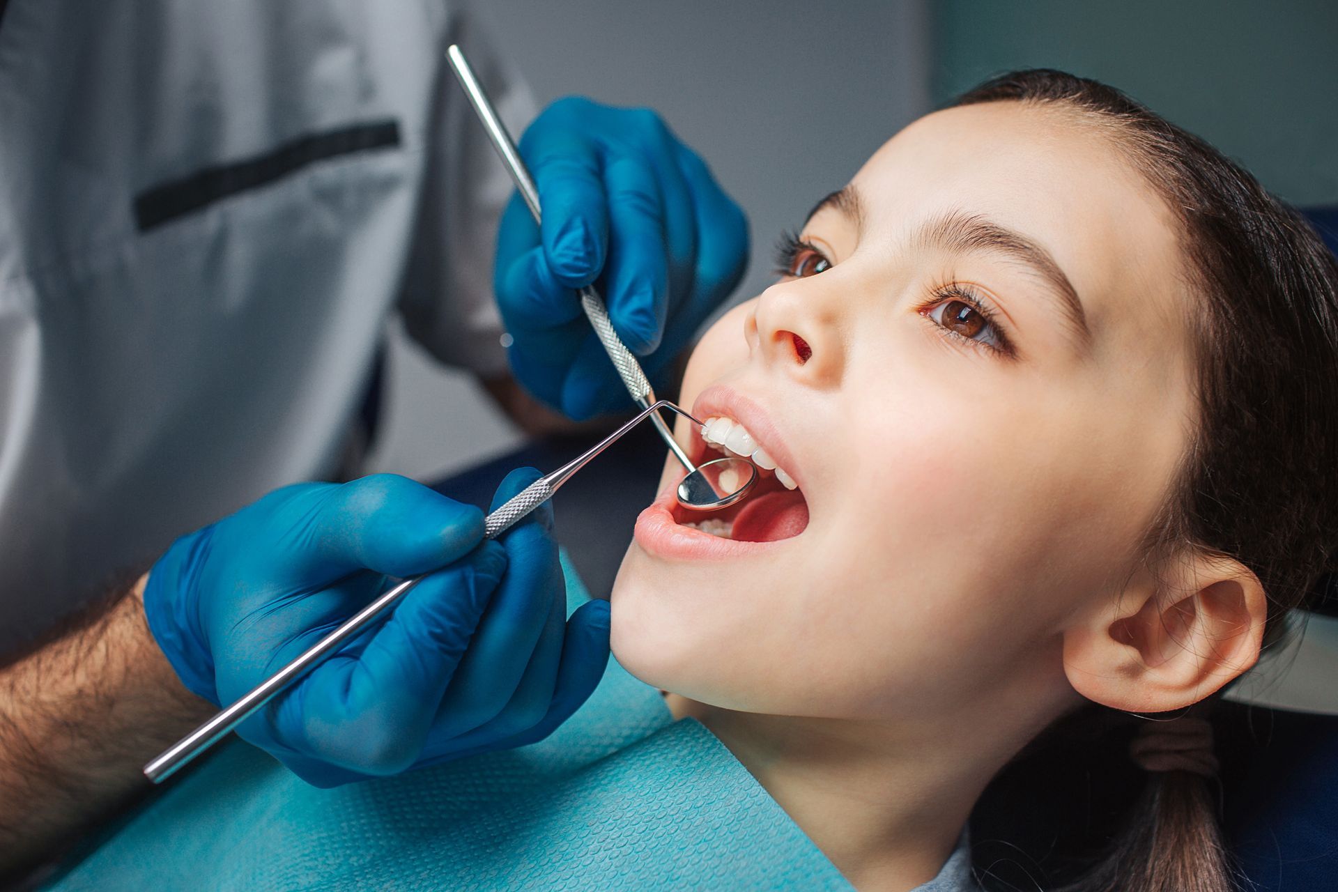 Un dentista, indossando guanti blu, esamina la bocca aperta di un paziente con specchietto e strumenti odontoiatrici in una clinica.