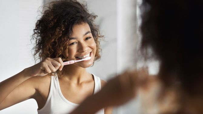 Una donna si lava i denti davanti allo specchio del bagno, sorridendo mentre indossa una canottiera bianca.