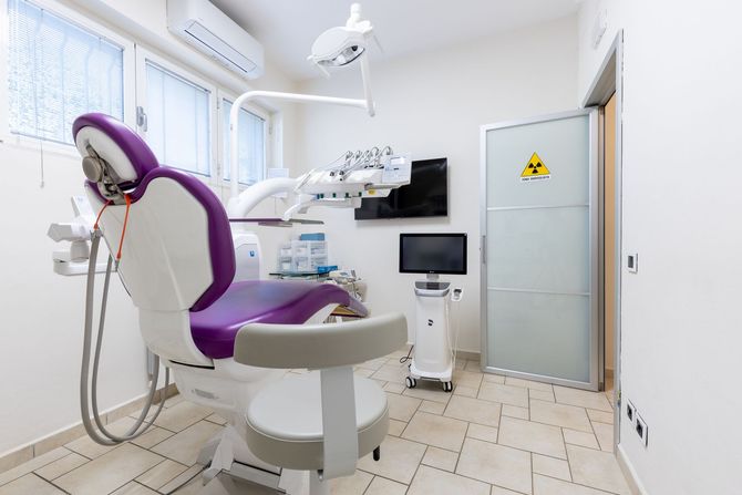 Clinica odontoiatrica moderna con poltrona per pazienti viola, attrezzature dentistiche, lavandino, monitor e porta satinata.
