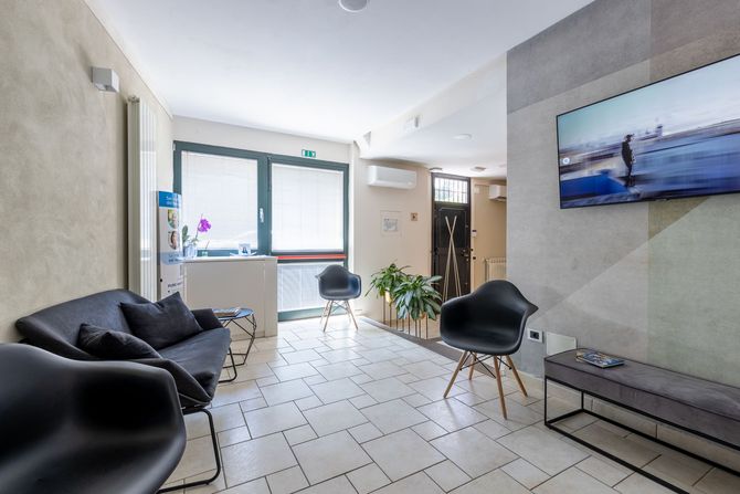 Sala d'attesa moderna con sedie nere, pavimento piastrellato, divano, pianta in vaso e una foto di paesaggio marino appesa alla parete.