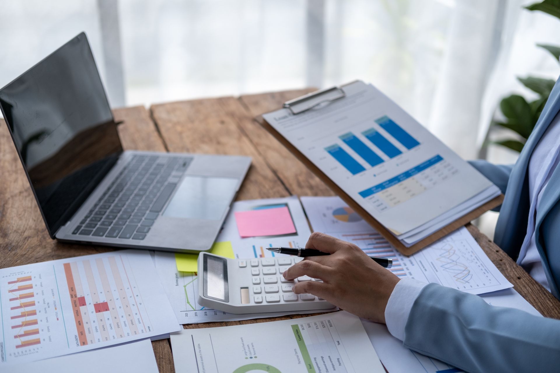 Person calculates finances with laptop, calculator, and charts on a wooden desk, wearing a blue suit.