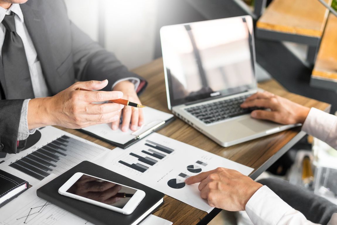Two businesspeople review financial documents at a desk with a laptop and smartphone.