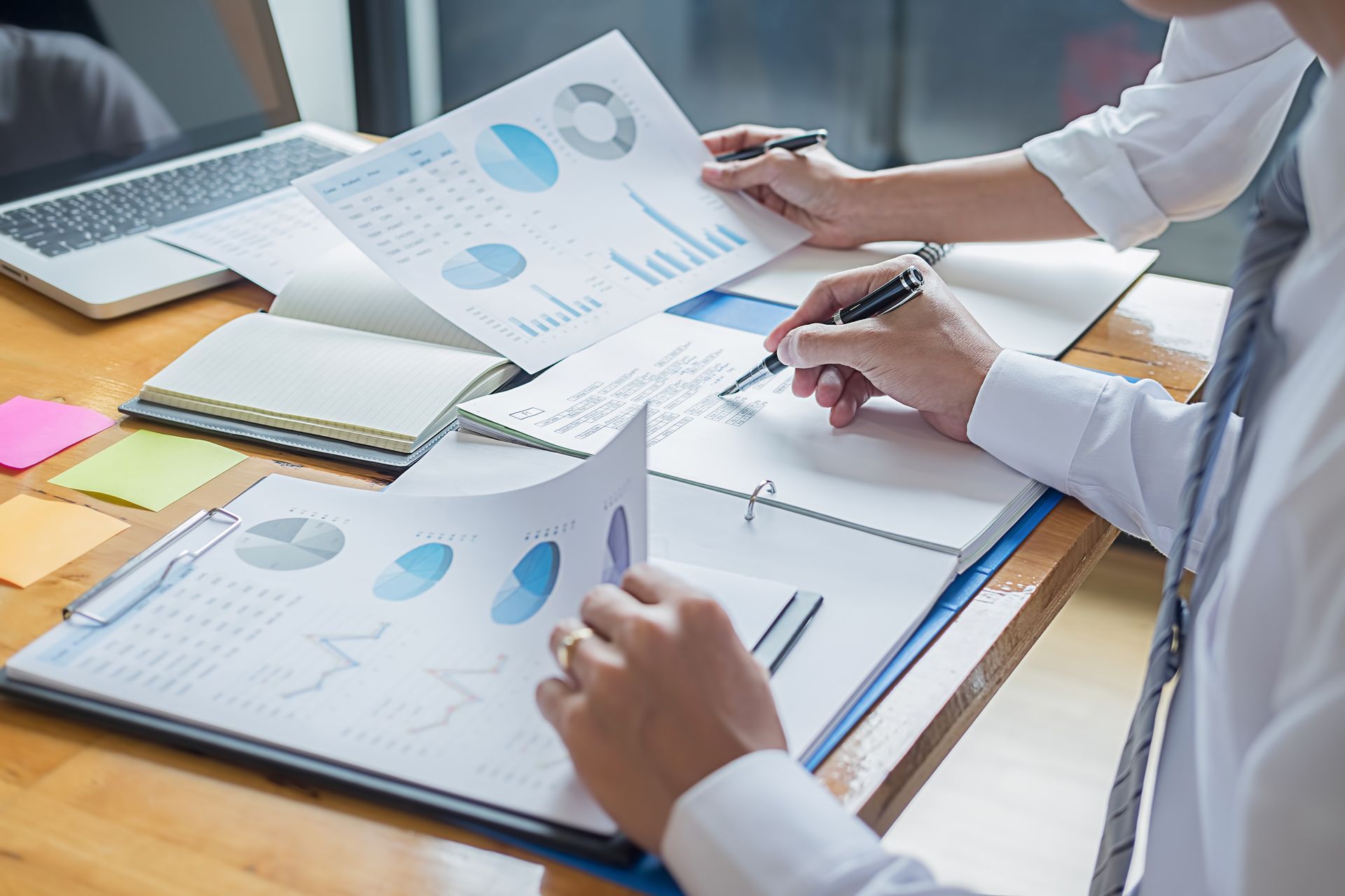 People reviewing charts and data on a desk; one points to a chart, the other writes notes.