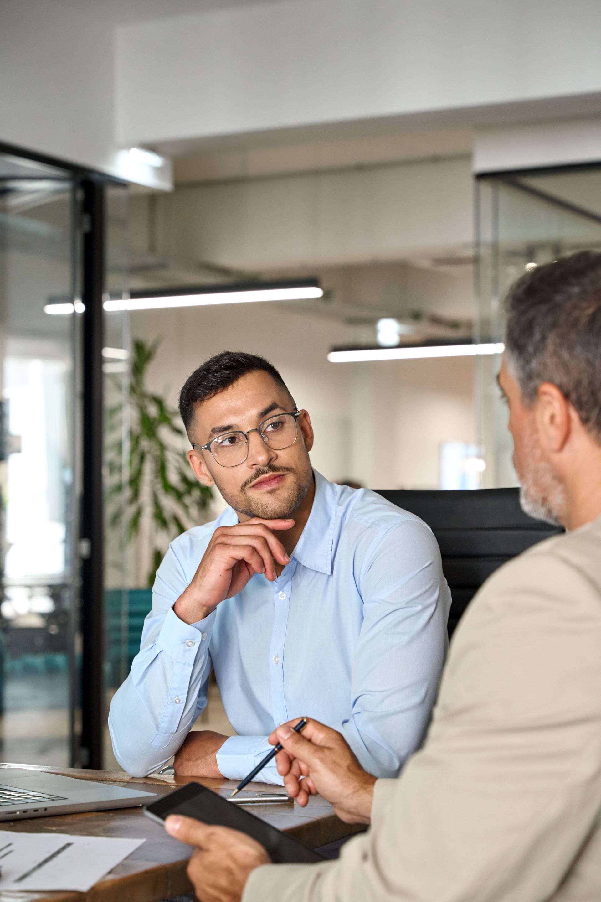 Two men in office setting, one in blue shirt, one in blazer, discussing, looking intently at each other.
