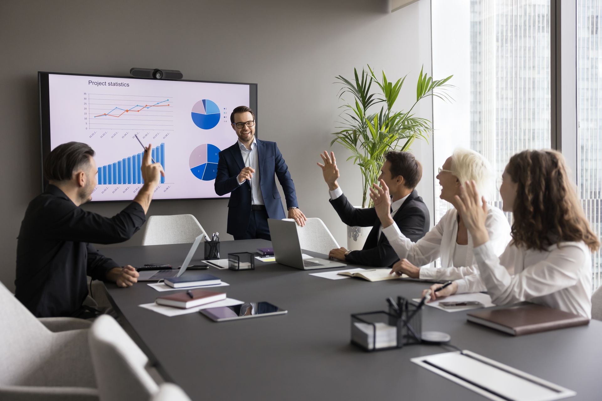 Business meeting: Man presenting data on screen, four people raising hands in response. Modern office setting.