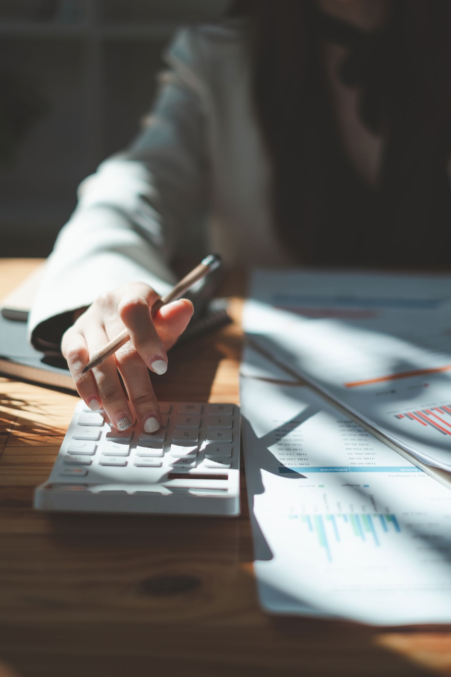 Person's hand using a calculator next to charts and graphs on a wooden desk.