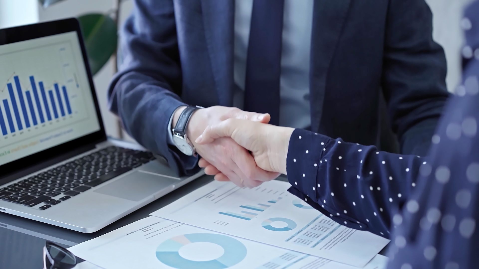 Two people in suits shaking hands over documents and a laptop displaying charts.
