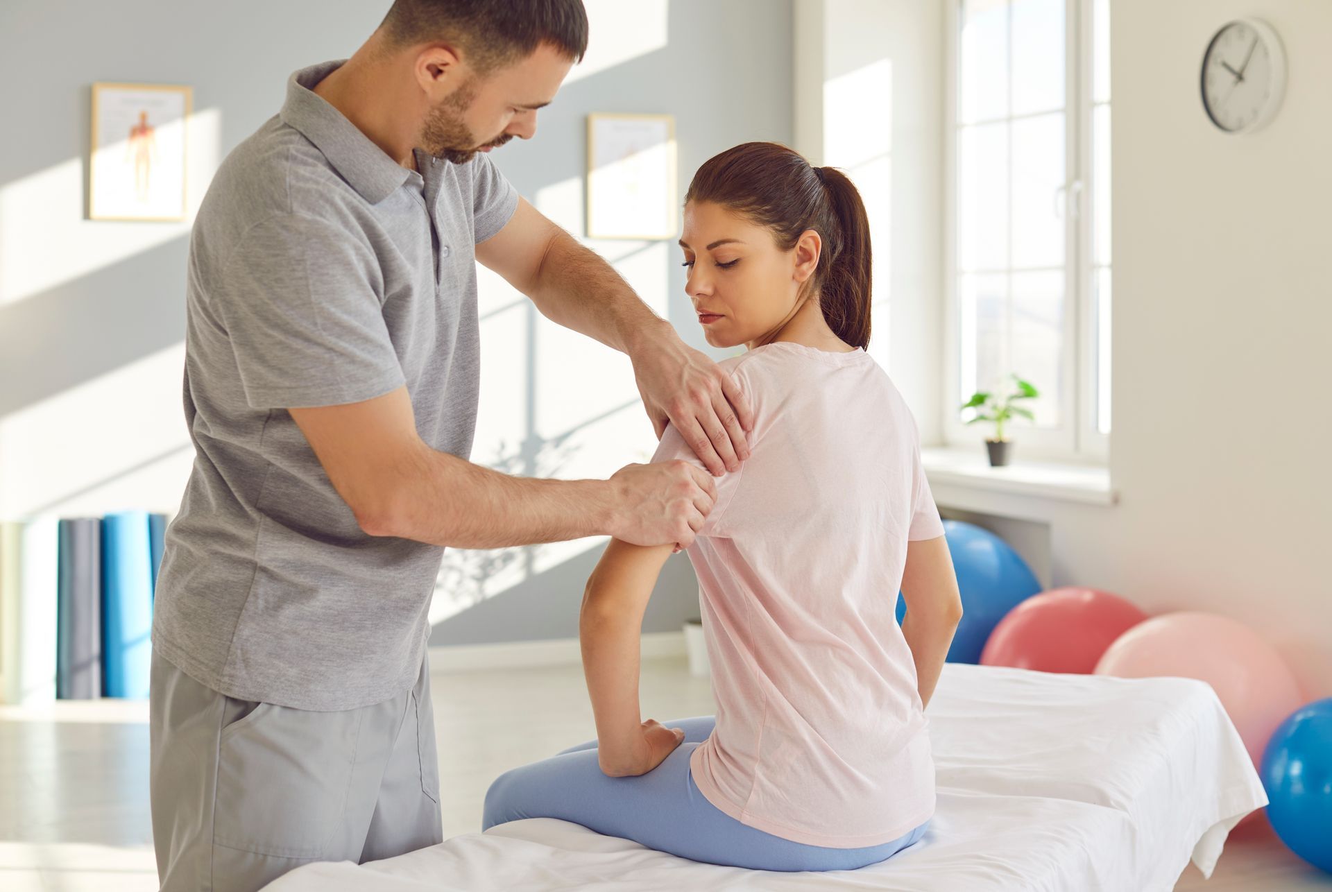 Chiropractor in scrubs gently assessing a seated patient's upper neck with a spine model on the desk Man, gently examining a seated woman's shoulder on a treatment table in a sunny chiropractor center.