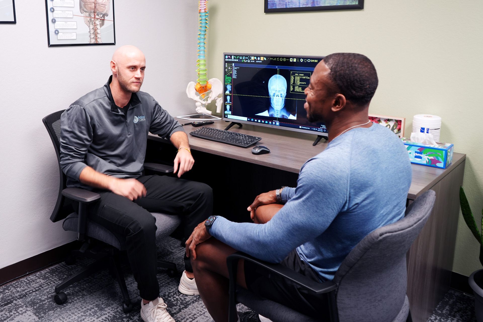 Two men seated, one behind a desk with a computer displaying a scan; discussing results in an office.