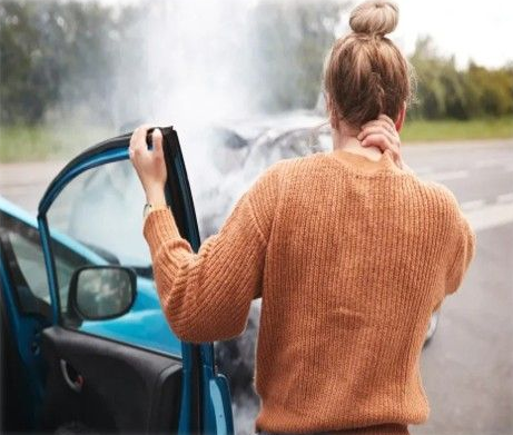 Woman with neck pain exits car after a collision; smoke rises from damaged vehicles.