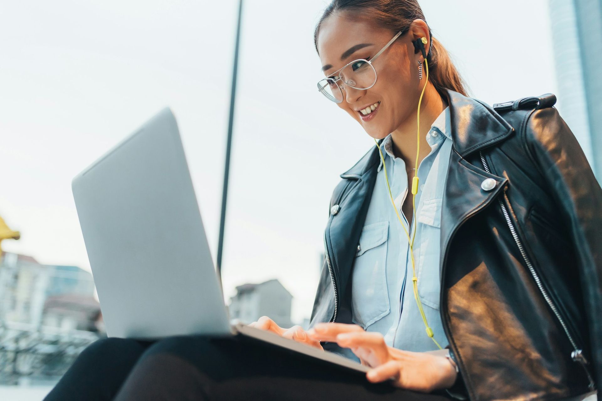 Woman with glasses and a black jacket uses a laptop and earbuds outdoors.