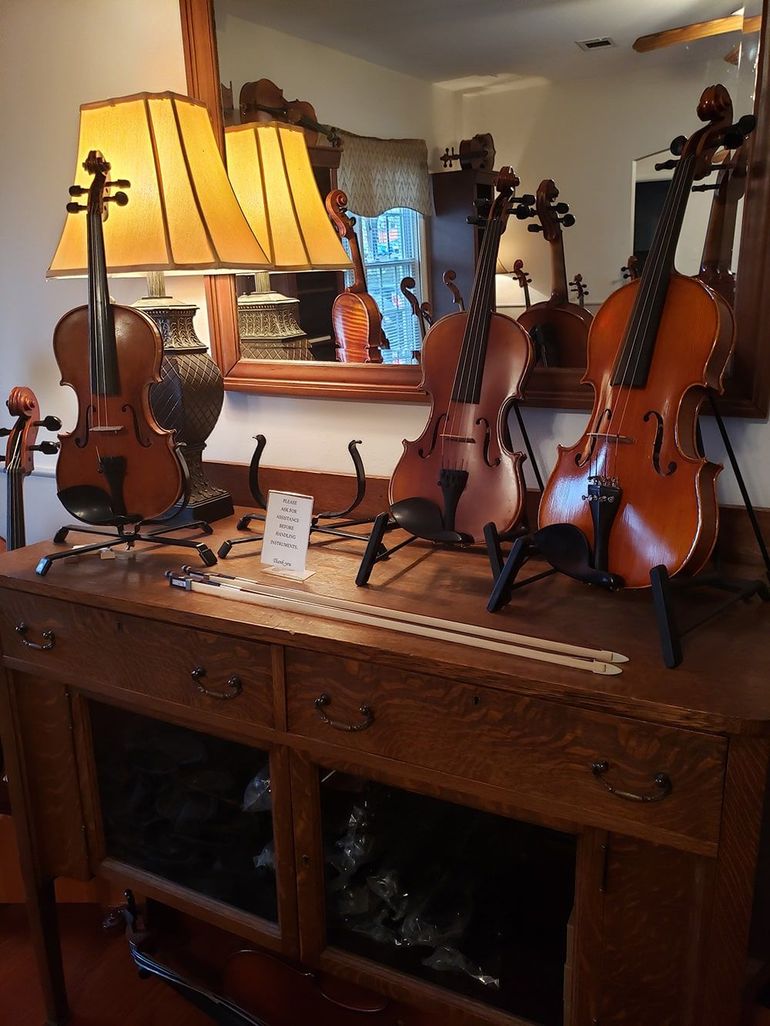 Three violins displayed on stands atop a wooden sideboard, with a table lamp and a mirror in the background.