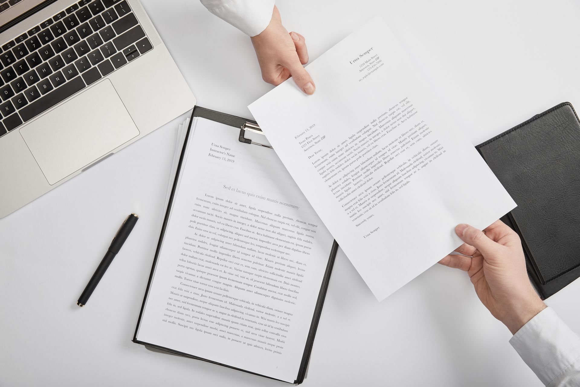 Mani che si scambiano un documento sopra una scrivania con computer portatile, blocco appunti, penna e fogli di carta.