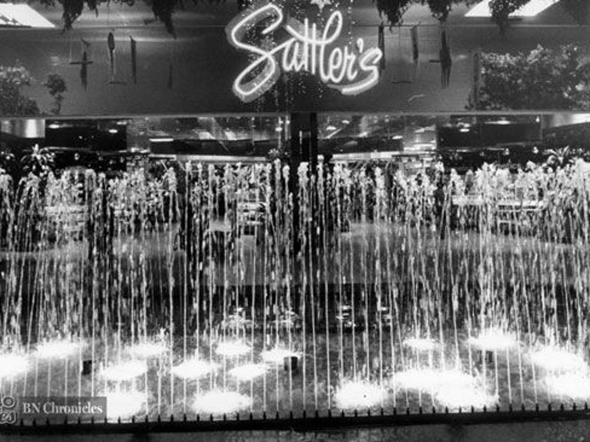 A fountain with multiple vertical jets of water in front of a storefront with a large, illuminated