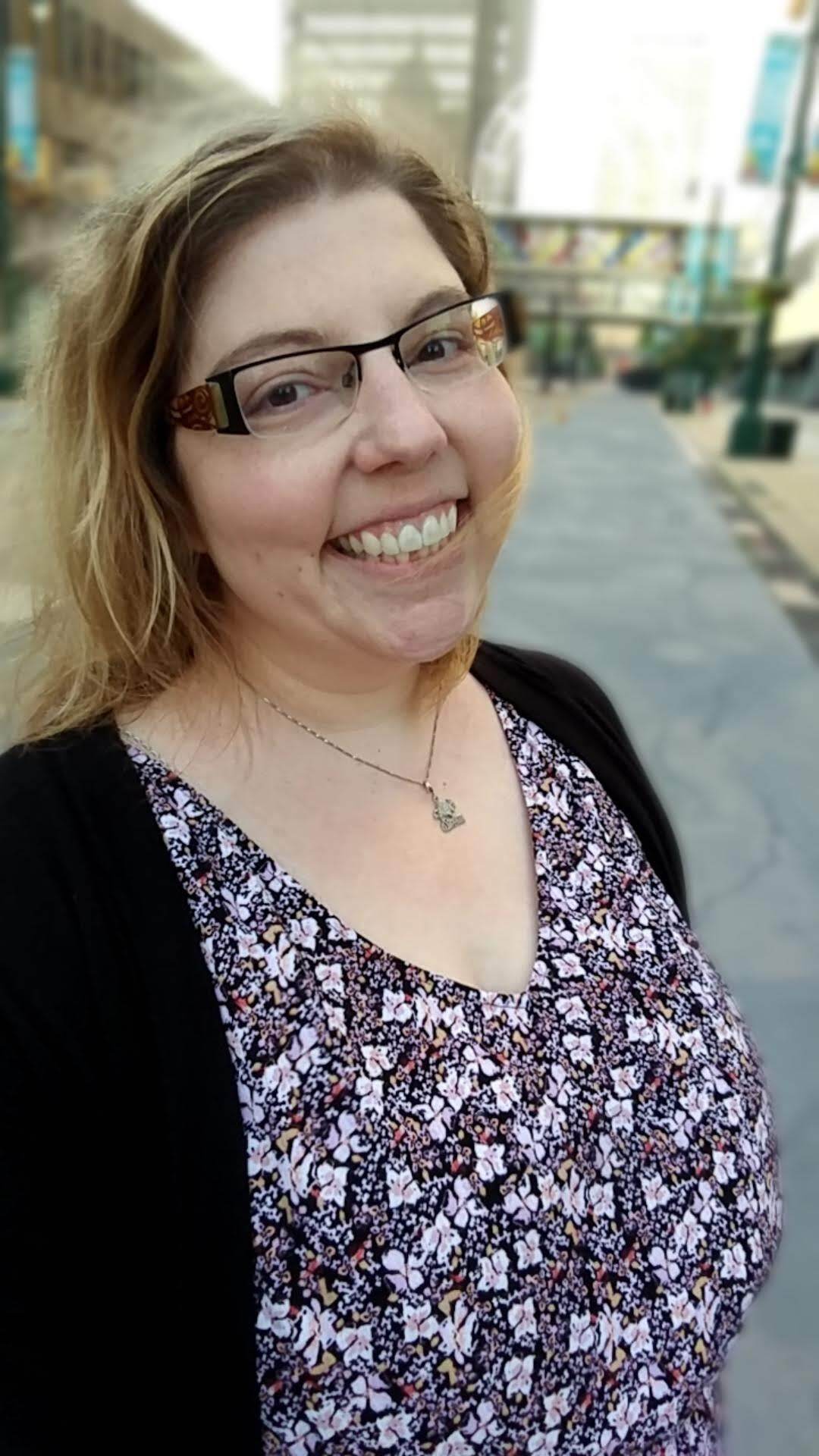 A smiling person wearing glasses, a patterned top, and a black cardigan outdoors on a paved path.