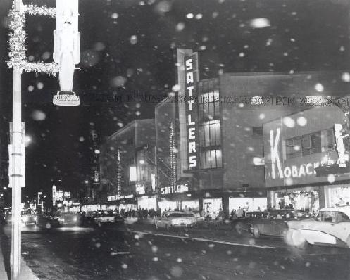 A black-and-white night view of a snow-covered street in Buffalo, featuring the illuminated Sattler's department store.