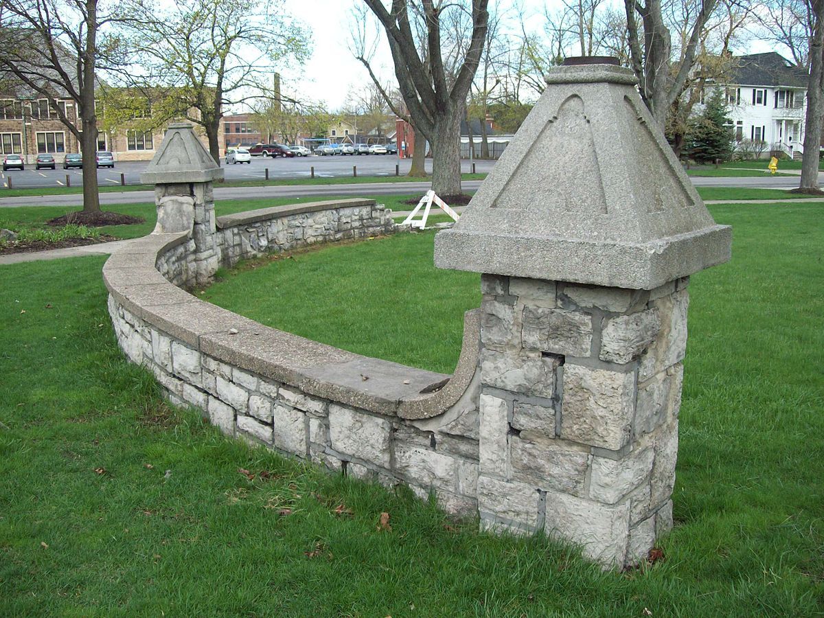 A curved stone wall with decorative stone pillars, located on a grassy lawn with trees and a parking lot in the background.