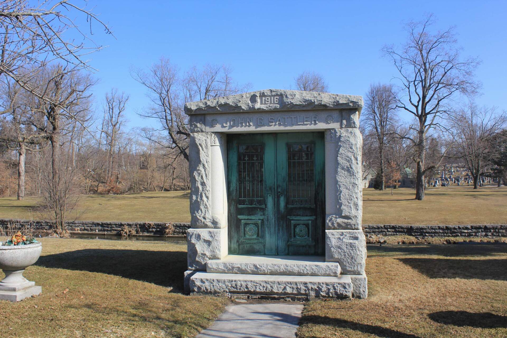 A stone mausoleum with ornate green metal doors stands on a grassy cemetery lawn under a clear blue sky.