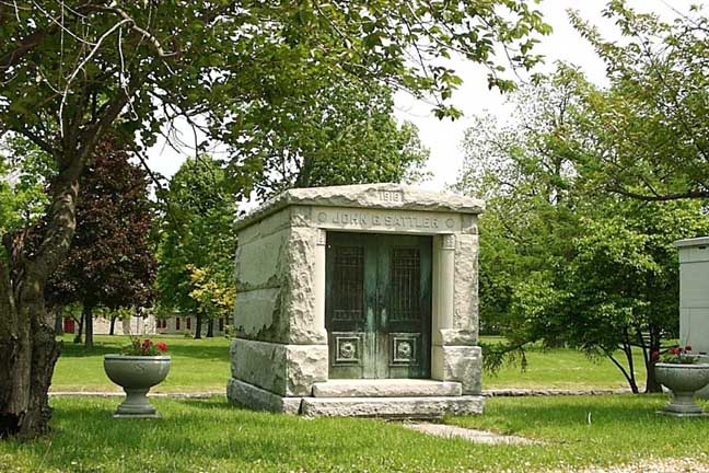 A stone mausoleum with metal doors sits in a grassy, tree-filled cemetery with decorative planters on either side.