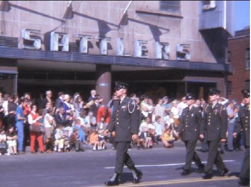 Uniformed marchers in a parade pass by a crowd in front of a building with a large
