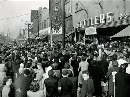 A dense crowd fills a city street, gathering outside a storefront with a large sign reading