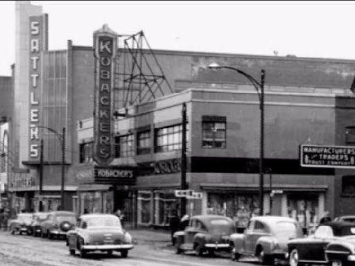 Black-and-white street-level view of a commercial district featuring prominent signs for Sattler’s and Kobacker’s stores.