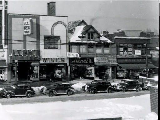 A street scene with snow-covered storefronts, including shops named Winch and Henri’s, with vintage cars parked in front.