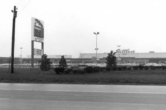 A black-and-white view of a Settler's store in a strip mall with a large signage pole in a gravel and paved lot.