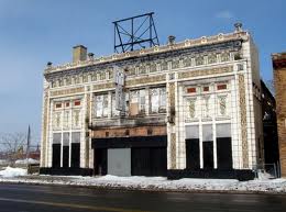 A two-story, weathered, white-tiled building with ornate details and a roof sign, standing beside a snowy road.